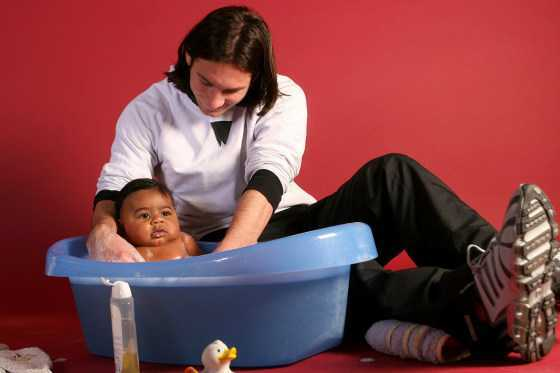 Messi in a white shirt sits on a red floor bathing a baby in a blue tub during a charity photoshoot, with a rubber duck beside them
